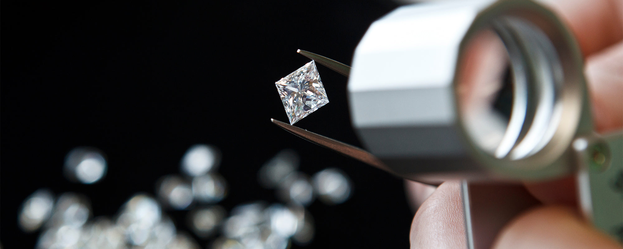 Close-up of a jeweller inspecting a princess-cut diamond with tweezers under a magnifying loupe. The diamond's precision-cut facets are clearly visible, reflecting light brilliantly. Several loose diamonds are blurred in the background, emphasising the main stone's clarity and quality. The image highlights the expert craftsmanship involved in diamond selection and quality control, making it ideal for showcasing the precision and attention to detail in fine jewellery creation.